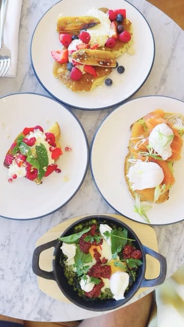 An overhead flat-lay photograph of a brunch spread featuring pancakes, French toast, green shakshuka, and smoked salmon avocado toast on a marble table.