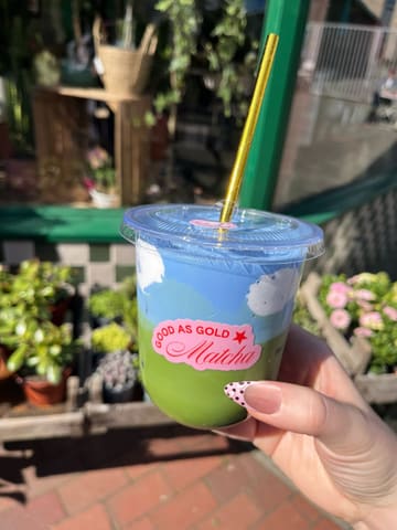 A close-up, eye-level shot of an iced matcha latte with cloud-shaped cold foam, held in a hand in front of a cafe storefront.