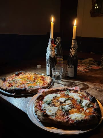 A moody, low-light shot of two pizzas served on metal trays at a table illuminated by two candles in wine bottles. The setting is intimate and atmospheric, typical of a pub or casual dining venue.