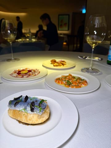 A high-quality, atmospheric shot of a fine dining table setting featuring multiple small, elegantly plated seafood dishes and wine glasses, capturing the sophisticated ambiance of a restaurant.