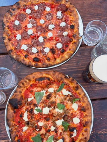 A high-quality, overhead flat-lay photograph of two pizzas served on a rustic wooden table, accompanied by glasses of water and a drink. The lighting is bright and natural, highlighting the textures of the crispy crusts and the fresh toppings. The composition is clean and inviting, effectively capturing the casual, social atmosphere of the venue.