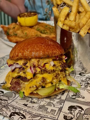 A close-up, appetizing shot of a double smash burger with melted cheese, pickles, and red onion, served on comic-book-themed paper with a side of fries in a metal cup.