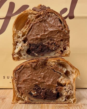 A high-quality, close-up cross-section shot of a chocolate-filled pastry, likely a cruffin, stacked on a wooden surface. The image effectively showcases the lamination of the pastry dough and the generous, rich chocolate filling, with the bakery's branded paper bag providing context in the background.