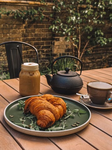 A high-quality, well-composed shot of a cafe table featuring a flaky croissant served on a bed of greens, accompanied by an iced coffee and a cast-iron teapot. The setting is an outdoor courtyard with a rustic brick wall background, evoking a relaxed, pleasant atmosphere.