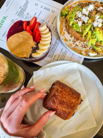 A bright, overhead flat-lay shot of a brunch spread at a cafe, featuring an acai bowl, avocado toast with feta, a pain au chocolat, and a matcha drink, captured in a clean, natural light setting.