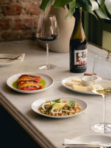 A high-quality, professional food photography shot featuring a spread of small plates and wine at a wine and vinyl bar. The composition is balanced, with a shallow depth of field drawing attention to the foreground dish, while the background elements like the wine bottle and greenery provide context and atmosphere.