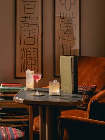 A sophisticated, atmospheric lifestyle shot showcasing two cocktails on a table within a stylish, book-filled hotel lounge setting. The image effectively captures the upscale, relaxed ambiance of the venue.