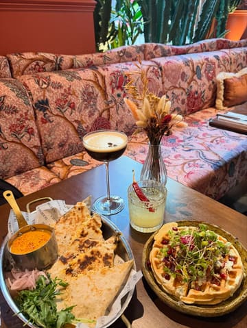 A high-quality, inviting shot of a brunch spread featuring quesadillas, a waffle dish, and two cocktails, set against a stylish, patterned booth background.
