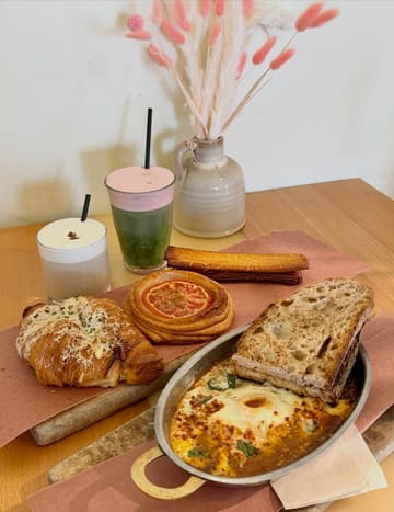 A high-quality, well-lit flat lay photograph showcasing a variety of brunch items from a bakery, including a tomato danish, a turkey croissant, baked eggs, and specialty drinks.