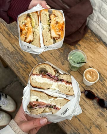 A high-quality, lifestyle-focused shot of two loaded bagels being held over a wooden table, accompanied by a matcha latte and a coffee. The image captures a casual, aesthetic brunch setting, likely at a bakery or cafe.