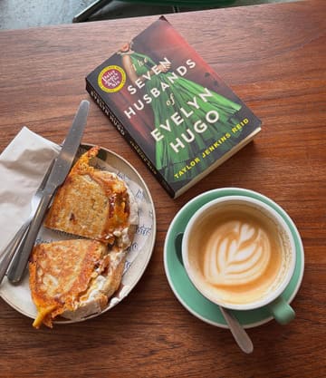 A flat-lay photograph taken from above featuring a toasted sandwich, a latte with latte art, and a copy of 'The Seven Husbands of Evelyn Hugo' on a wooden table, capturing a cozy cafe reading vibe.