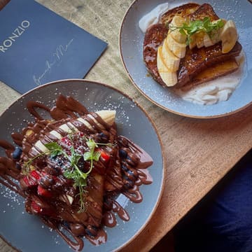 A top-down, overhead shot of two plates of French toast served at a cafe, featuring one chocolate-drizzled version with berries and one with banana and cream. The image is taken on a wooden table with a menu visible in the corner.