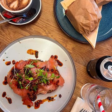 A well-composed, overhead flat-lay photograph of a brunch spread, featuring smoked salmon on toast topped with chilli crisp, accompanied by a coffee and a wrapped sandwich on a wooden table.