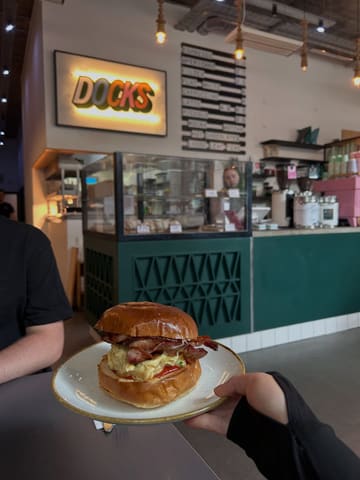 A casual, first-person perspective shot of a bacon and egg brioche bun held in front of a cafe counter. The image captures the atmosphere of the venue, with the menu board and coffee equipment visible in the background.