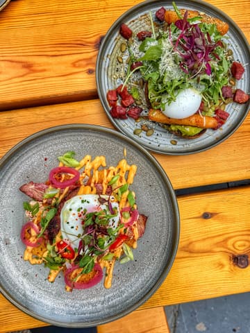 An overhead, flat-lay shot of two brunch dishes served on a wooden table. The top plate features avocado toast topped with a poached egg, microgreens, and what appears to be chorizo or sun-dried tomatoes. The bottom plate features a potato-based dish, likely hash browns, topped with bacon, a poached egg, spicy mayo, and pickled red onions.