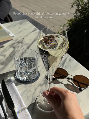 A close-up shot of a glass of sparkling wine held in a hand on an outdoor marble table, accompanied by sunglasses and a glass of water, capturing a sunny, relaxed atmosphere.