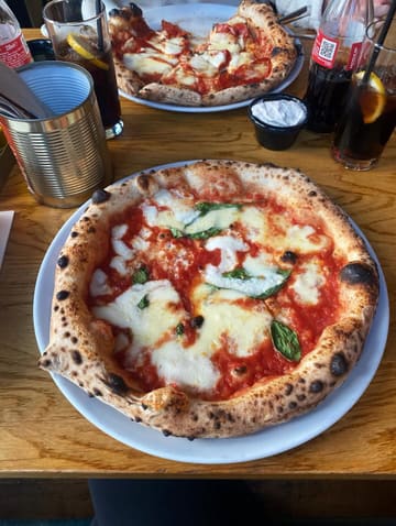 A top-down, slightly angled shot of a freshly baked Neapolitan-style pizza on a wooden table, with a second pizza and drinks visible in the background, capturing a casual dining experience.