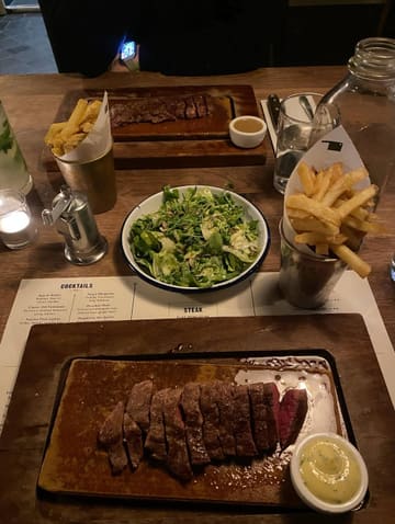 A top-down shot of a steak dinner at a restaurant, featuring two steak boards, fries, and a salad. The lighting is dim and atmospheric, typical of a steakhouse setting.