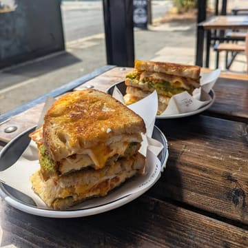A close-up, appetizing shot of two toasted sandwiches served on parchment-lined plates at an outdoor cafe table. The image highlights the textures of the sourdough, melted cheese, and fresh ingredients, effectively capturing a casual brunch atmosphere.