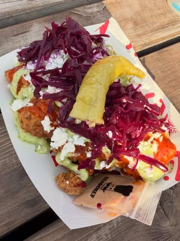 A close-up, high-angle shot of a portion of chicken wings topped with red cabbage, feta cheese, sauce, and a whole pickled pepper, served in a paper tray on a wooden table.