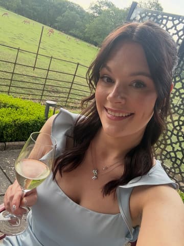 A high-quality, personal selfie taken at an outdoor venue featuring a glass of wine and a scenic deer park in the background, capturing a celebratory atmosphere.