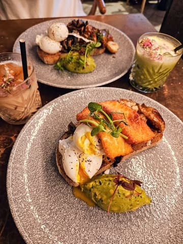 A top-down, overhead shot of a brunch spread featuring two plates of food and two drinks. The main focus is on a plate with poached eggs, halloumi, avocado, and mushrooms, while a second plate is partially visible at the top of the frame.