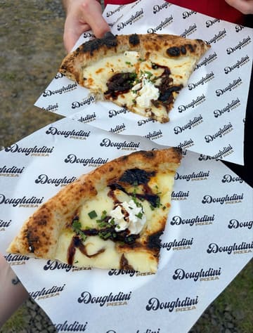 A high-quality, overhead shot of two pizza slices held by a person, served on branded 'Doughdini Pizza' paper. The pizza features a charred crust, melted cheese, and a balsamic glaze drizzle, presenting a fresh and appetizing street food aesthetic.