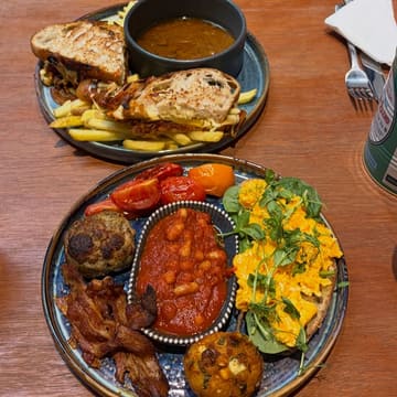 A high-quality, overhead shot of a brunch spread featuring a short rib grilled toastie with a side of jus and a 'big breakfast' plate containing eggs, bacon, sausage, beans, and a unique hash brown.