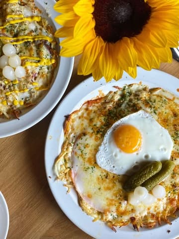 A bright, overhead flat-lay shot of two Swiss-style röstis served on white plates, garnished with pickles and a fried egg, accompanied by a large sunflower.