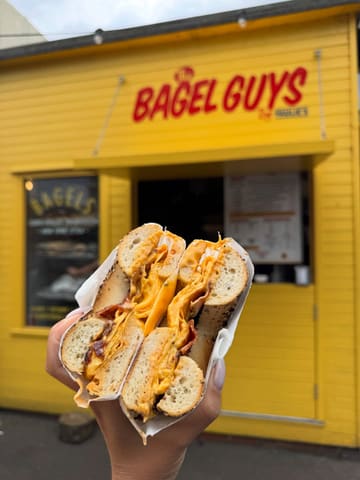 A close-up, handheld shot of a bacon, egg, and cheese bagel held in front of a bright yellow 'The Bagel Guys' market stall. The focus is sharp on the textures of the bagel and the melted cheese, with the background softly blurred to keep attention on the food.