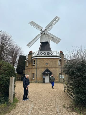 A candid, wide-angle snapshot of an elderly man and a woman standing in front of the Wimbledon Windmill museum on a grey, overcast day. The image functions as a personal travel memory rather than a polished marketing asset for the venue.