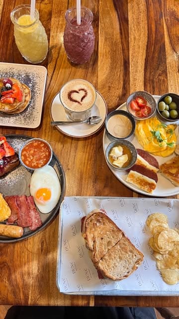 A high-angle, flat-lay shot of a brunch spread featuring pancakes, a full English-style breakfast, a sandwich with chips, and various drinks on a wooden table.