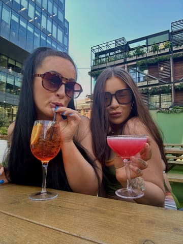 A photo of two women posing with cocktails at an outdoor venue, capturing a social, sunny atmosphere.