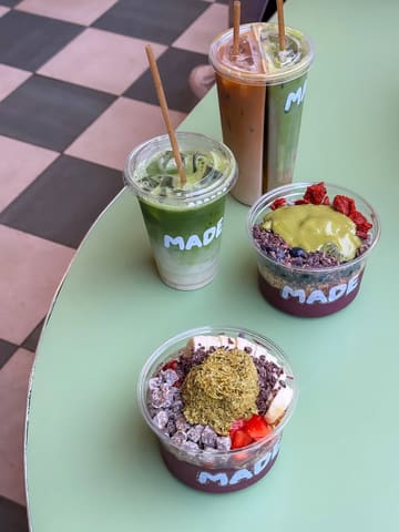 A bright, overhead shot of three drinks and two acai bowls arranged on a green table, showcasing the vibrant colors and toppings of the food and drink items at a cafe.