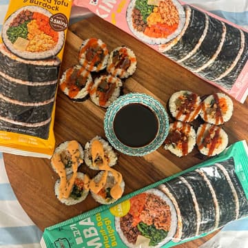 A bright, overhead flat-lay photograph showcasing frozen kimbap products. The image displays the product packaging alongside the prepared, sliced kimbap rolls, which are garnished with a sauce drizzle and accompanied by a small bowl of soy sauce on a wooden board.