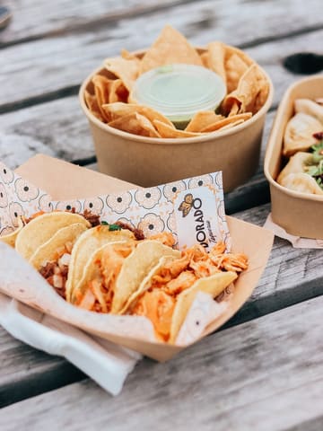 A high-angle, natural light shot of a tray of tacos from a street food market, set on a wooden picnic table. The primary subject is the tacos in the foreground, with a bowl of tortilla chips and a container of dumplings visible in the background.