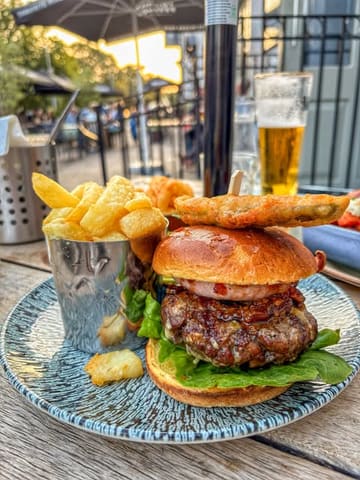 A high-quality, well-lit shot of a pub-style beef burger served with chips, captured during golden hour on an outdoor terrace. The image effectively conveys a relaxed, enjoyable dining experience.