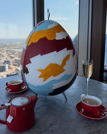 A high-angle shot of an afternoon tea service set against a panoramic city skyline view from a high-rise venue. The table features a large, artistically painted egg-shaped centerpiece, a red teapot, matching teacups, and a glass of champagne, evoking a sophisticated and scenic dining experience.