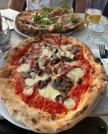 A casual, top-down shot of two Neapolitan-style pizzas on a table. The foreground pizza is the primary focus, displaying toppings such as mushrooms, olives, and ham, while a second pizza and a glass of beer are visible in the background.