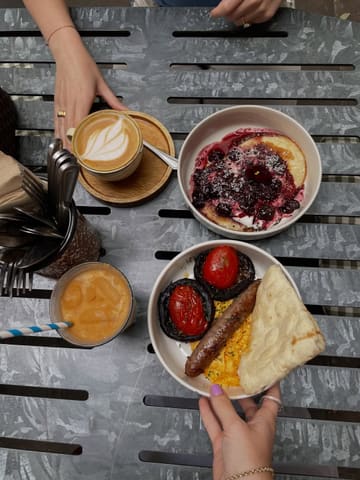 An overhead flat-lay shot of a brunch spread featuring pancakes with berry compote, a cooked breakfast plate with sausage, mushrooms, tomatoes, and flatbread, and a latte.