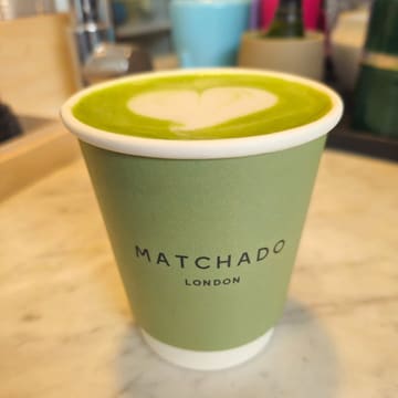 A close-up, eye-level shot of a matcha latte in a branded paper cup, featuring latte art, placed on a marble surface.