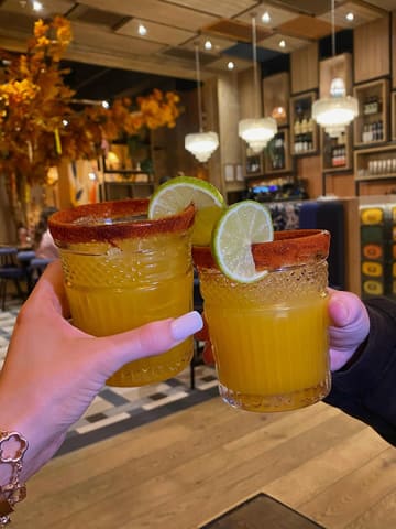 A first-person perspective shot of two people holding up mango margaritas in a restaurant setting, capturing a social 'cheers' moment.