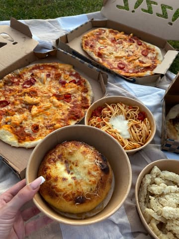 A top-down, flat-lay style photograph of a takeaway picnic spread on a blanket, featuring two pizzas, a garlic and mozzarella bombe, a bowl of spaghetti, and a bowl of calamari. The lighting is natural and warm, consistent with an outdoor sunset setting.