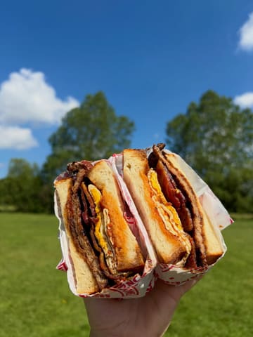 A bright, high-quality close-up shot of a breakfast sandwich held against a clear blue sky and green field background. The natural lighting is excellent, effectively highlighting the textures of the toasted bread, sausage, bacon, and egg.