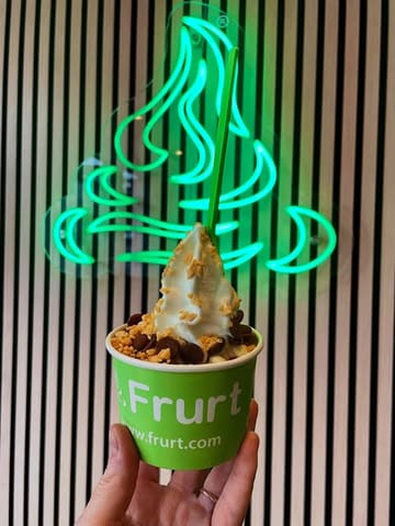 A casual, user-generated style photo of a frozen yogurt cup held in front of a neon sign at a Frurt venue.