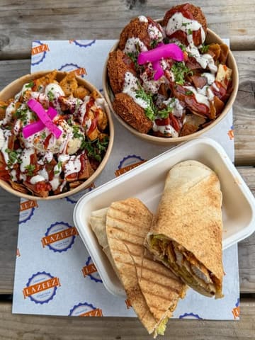 A top-down, overhead shot of three Lebanese street food dishes—a chicken shawarma wrap, a chicken protein box, and a falafel street box—served in cardboard containers on branded paper at an outdoor wooden table.