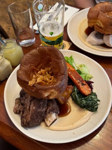 A high-quality, appetizing shot of a classic Sunday roast dinner at a pub, featuring a large Yorkshire pudding, beef, and vegetables, accompanied by a pint of beer.