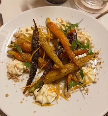 A top-down, well-composed shot of a roasted carrot dish featuring whipped feta and a crumble topping, served in a modern restaurant setting.