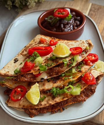 A high-quality, appetizing close-up shot of homemade pulled pork quesadillas served with a side of black bean dip. The dish is garnished with fresh herbs, sliced chili, and lime wedges, presented on a simple, clean plate.