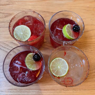A top-down, well-lit photograph of four different margaritas arranged on a wooden table. The drinks are garnished with lime wheels, cherries, and strawberries, with some featuring a chili-salt rim, presenting a vibrant and inviting selection of cocktails.